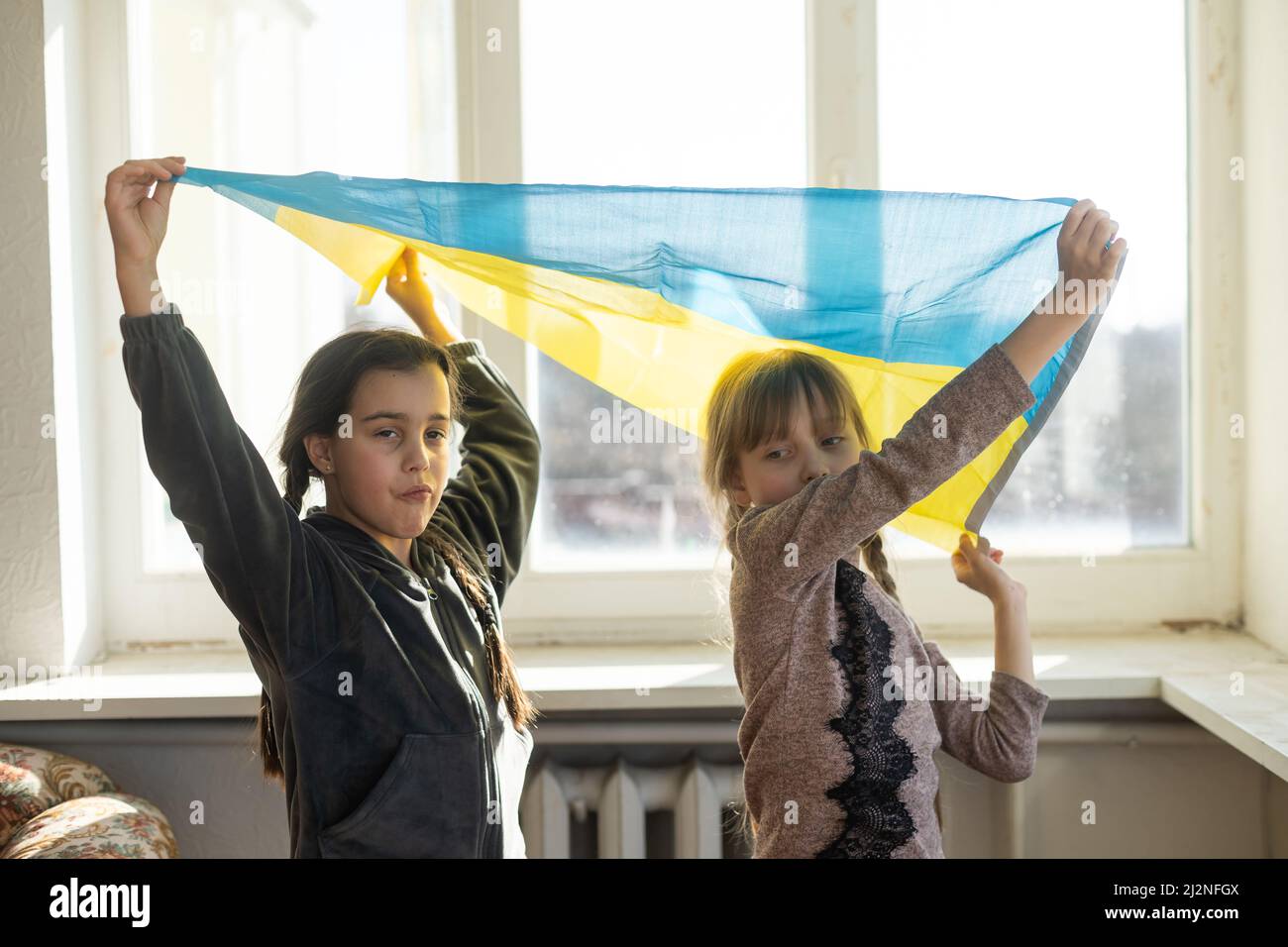 Kleine Mädchen mit ukrainischer Flagge vor einem Fenster. Die kleinen Mädchen winken die Nationalflagge, während sie für den Frieden beten Stockfoto