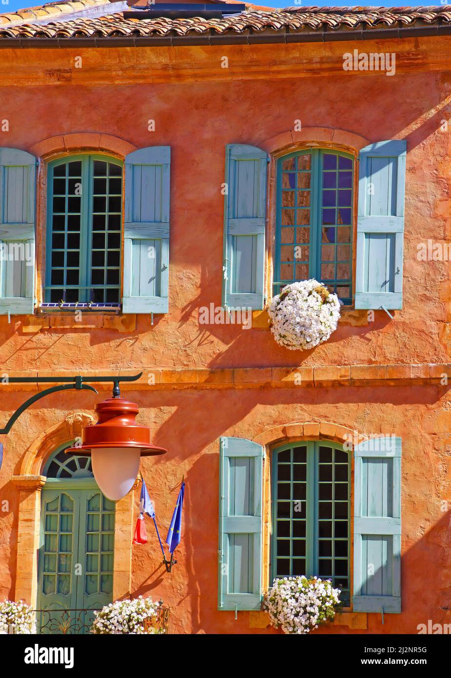 Blick auf die traditionelle bunte ockerfarbene Fassade eines Natursteinhauses aus Stein, Blumenarrangements, alte Fensterläden aus Holz - Rathaus von Roussillon, Frankreich Stockfoto