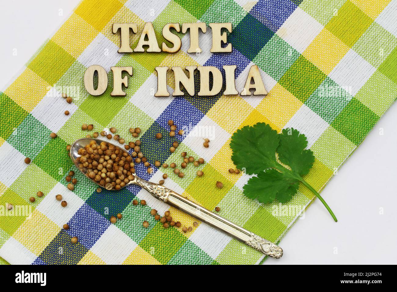 Taste of India mit Holzbuchstaben auf bunt kariertem Tuch mit frischen Korianderblättern und Koriandersamen auf Vintage-Löffel geschrieben Stockfoto