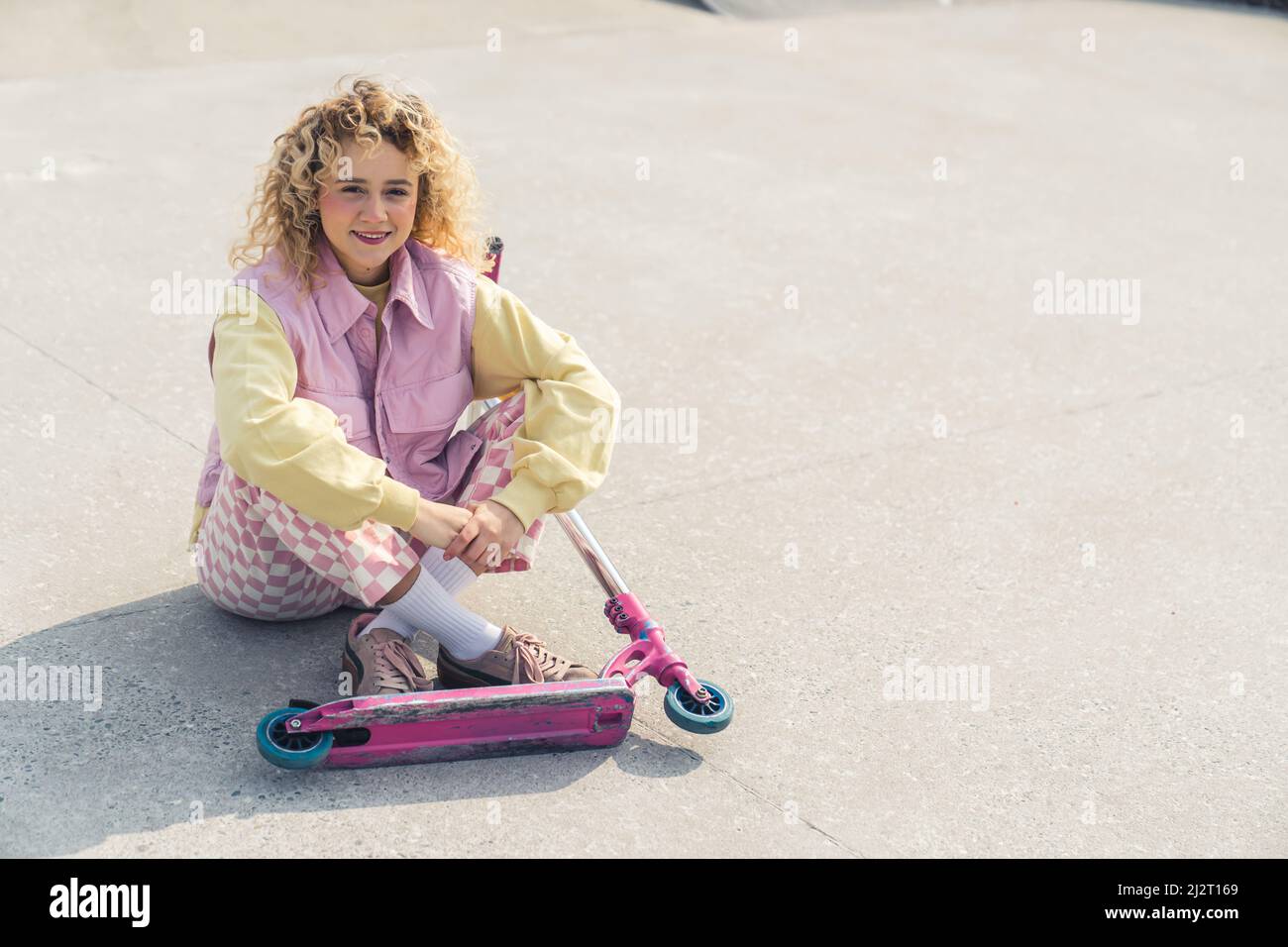 Die attraktive junge, frisch gesichtige blonde Hipster-Frau sitzt lächelnd auf dem Sportplatz, die Beine gekreuzt, der rosarote Roller vor ihrem Blick auf die Kamera voller Aufnahmen. Hochwertige Fotos Stockfoto