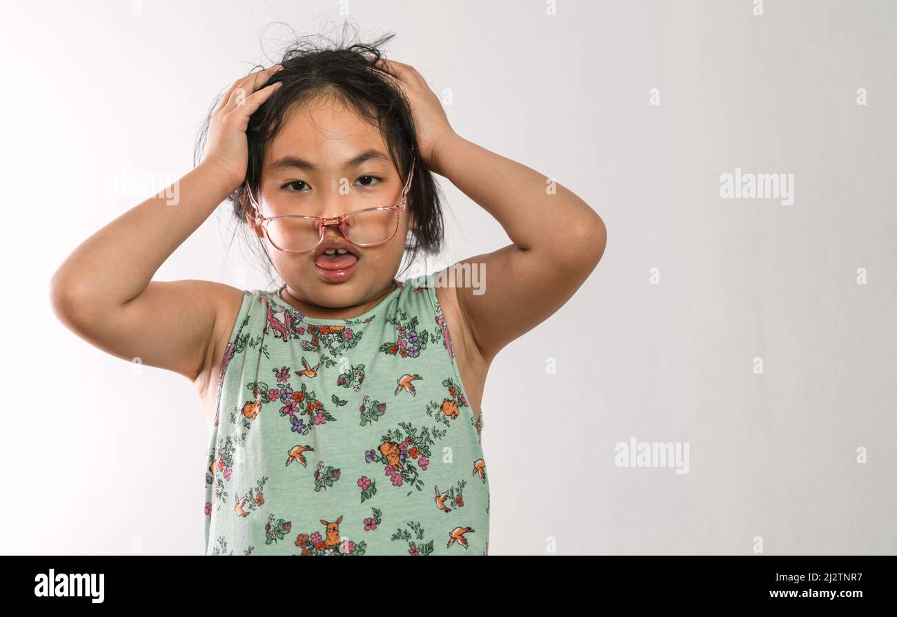 Porträt eines verwirrten asiatischen kleinen Mädchens, beide Hand auf Kopf, heruntergeklappte Brille auf dem Gesicht, unordentliches Haar. Das Konzept der Kindererziehung. Stockfoto