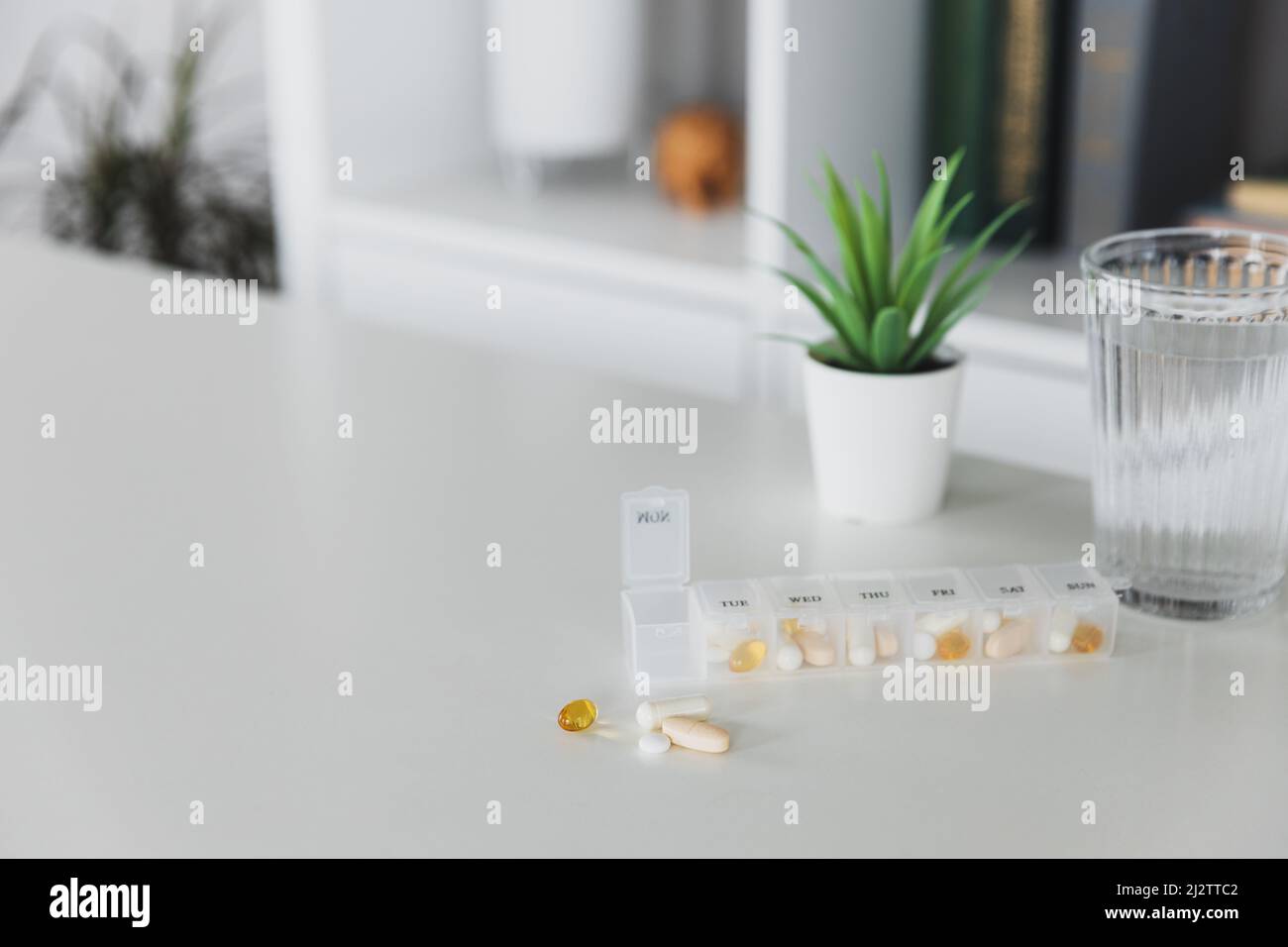 Nahaufnahme der medizinischen Pille Box mit Dosen von Tabletten für den täglichen Nehmen Sie ein Medikament zur Behandlung, die Krankheit zu heilen. Glas Tasse Wasser auf dem Tisch, grauer Hintergrund. Grüner Blumentopf oder Pflanze. Helathy Lifestyle Stockfoto