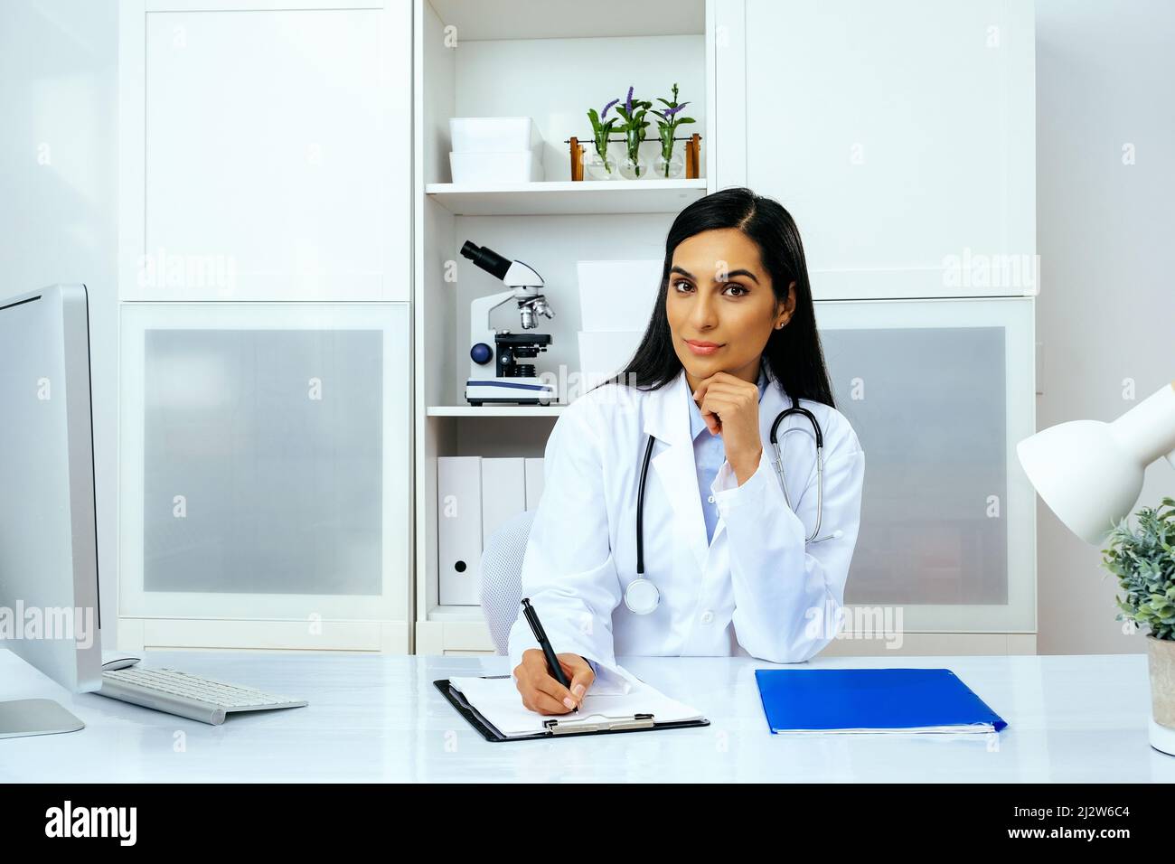 Porträt einer schönen jungen Ärztin in weißer Laborkittel-Uniform, die am Schreibtisch in ihrer modernen Büro-Gesundheitsbranche sitzt Stockfoto