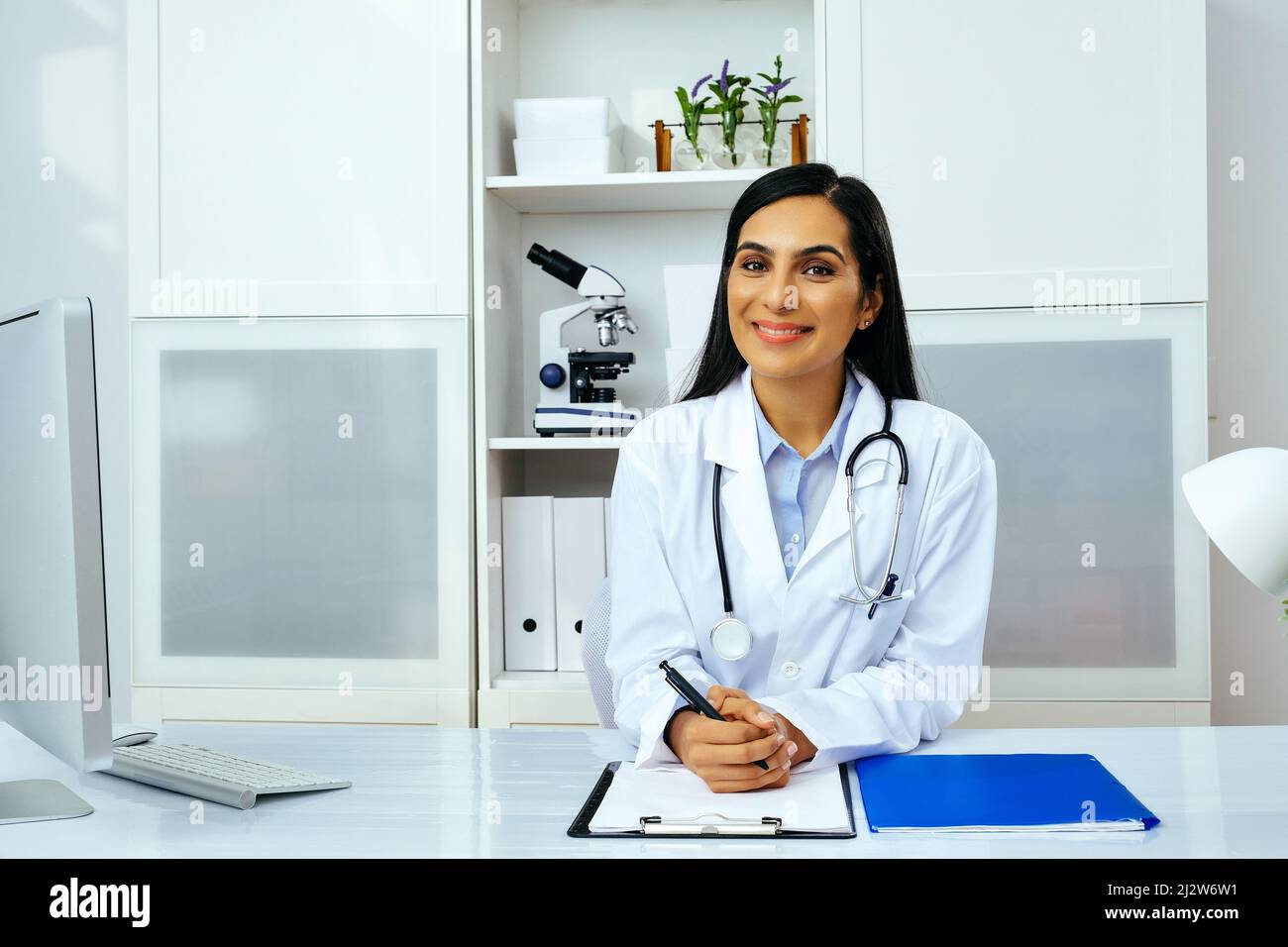 Porträt einer schönen jungen Ärztin in weißer Laborkittel-Uniform, die am Schreibtisch in ihrer modernen Büro-Gesundheitsbranche sitzt Stockfoto