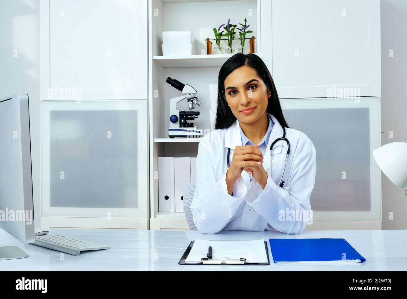 Porträt einer schönen jungen Ärztin in weißer Laborkittel-Uniform, die am Schreibtisch in ihrer modernen Büro-Gesundheitsbranche sitzt Stockfoto