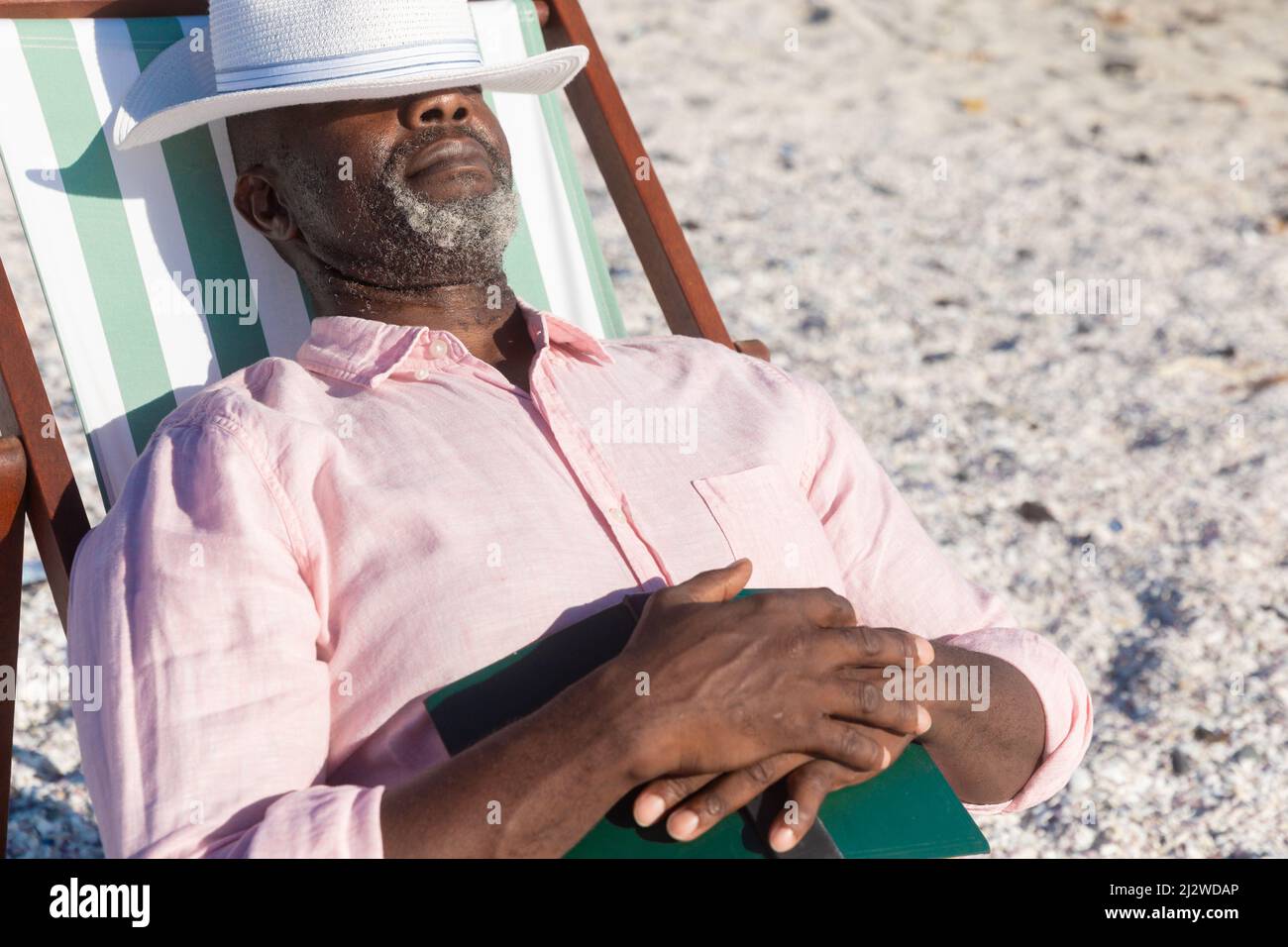 Pensionierter afroamerikanischer älterer Mann, der mit Buch und Hut über den Augen auf einem Klappstuhl am Strand schlief Stockfoto