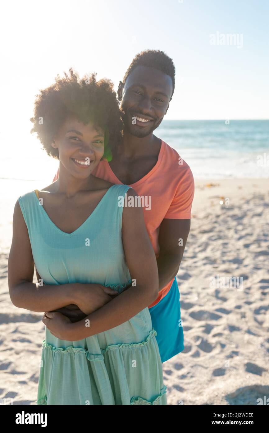 Hintergrundbeleuchtetes Porträt eines lächelnden afroamerikanischen Mannes, der seine Freundin von hinten am Strand umarmt Stockfoto