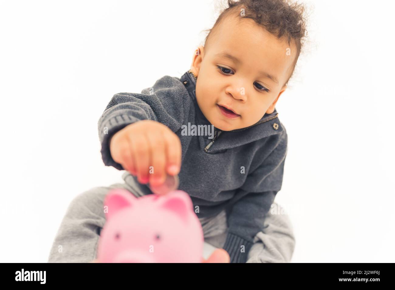 Süßes afroamerikanisches Kleinkind, das auf dem Boden sitzt und eine Münze in das rosafarbene Sparschwein-Studio-Shot mit mittlerer Vollaufnahme mit weißem Hintergrund kopiert. Hochwertige Fotos Stockfoto