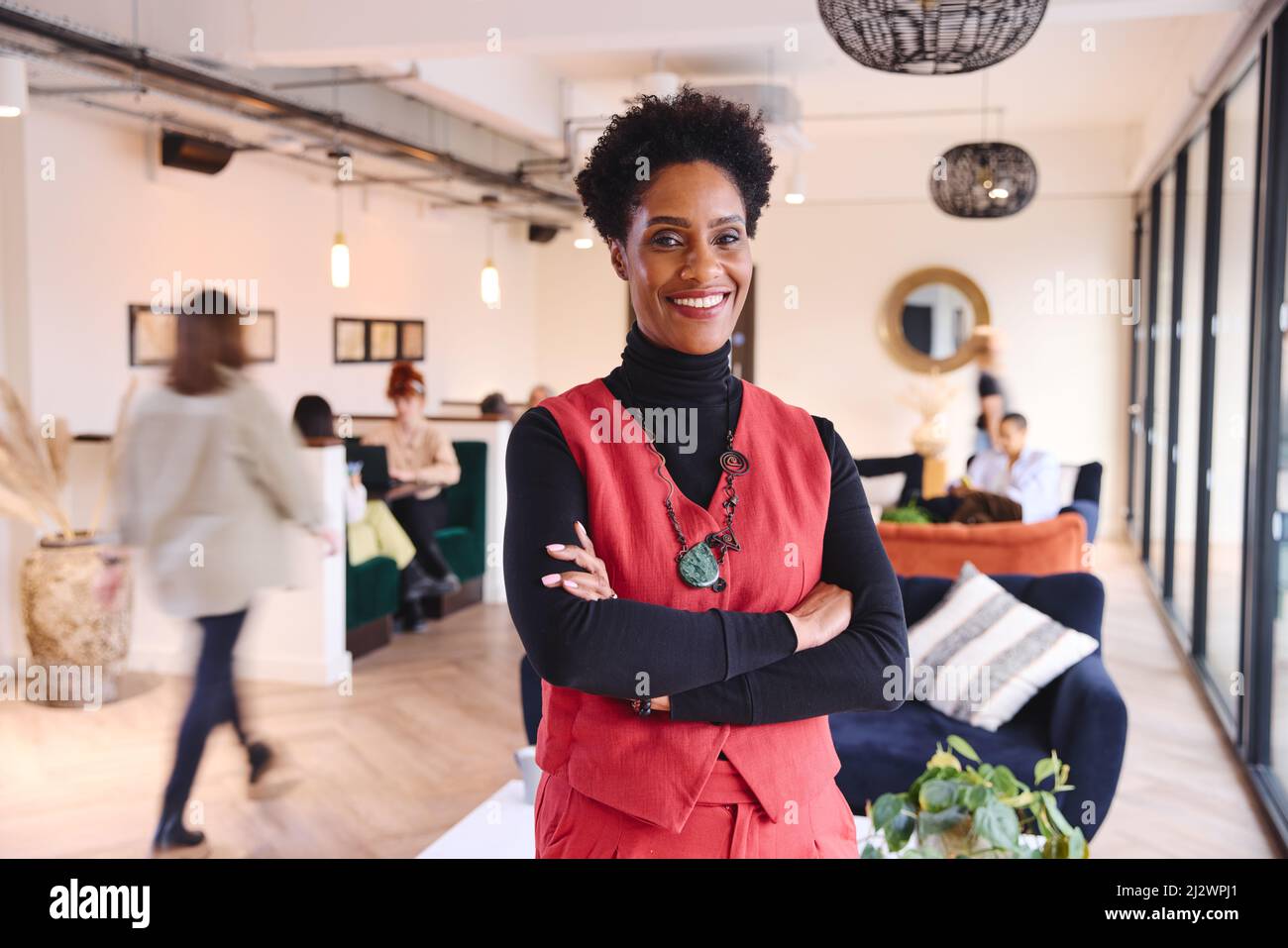 Porträt einer selbstbewussten, reifen schwarzen Geschäftsfrau mit kurzen schwarzen Haaren, die lächelt und mit gefalteten Armen im Coworking Space auf die Kamera blickt Stockfoto