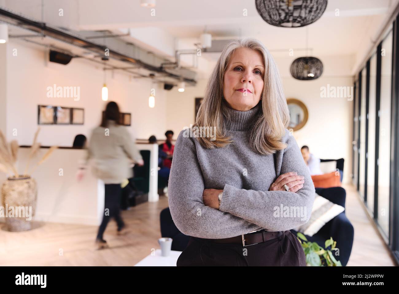 Porträt einer selbstbewussten, weißen Geschäftsfrau mit langen grauen Haaren, die die Kamera mit gefalteten Armen im Coworking Space anschaut Stockfoto
