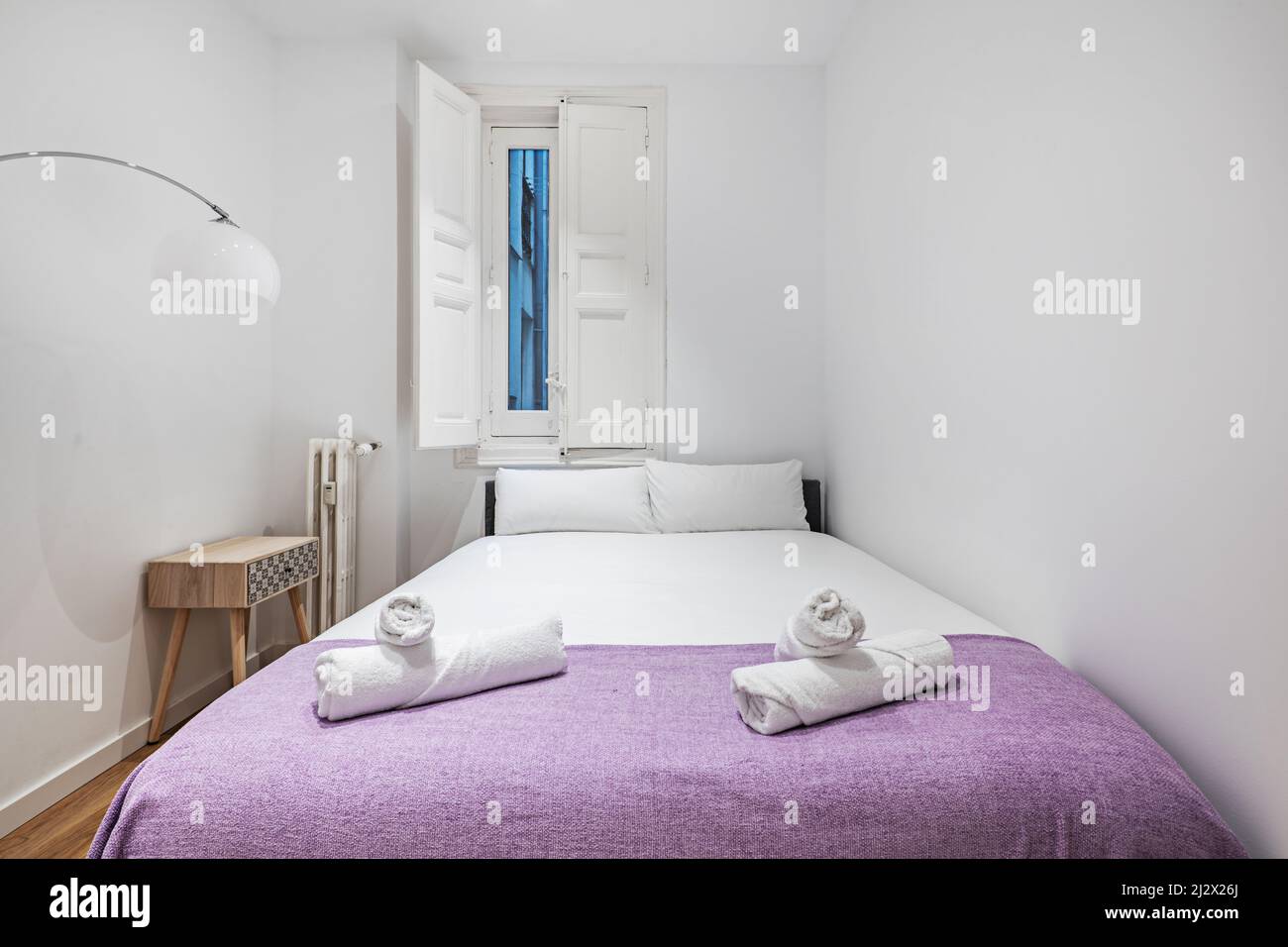 Schlafzimmer mit Doppelbett mit violetter Decke auf dem Bett, gusseisernem Heizkörper und Fenster mit weißen Holzfenstern Stockfoto