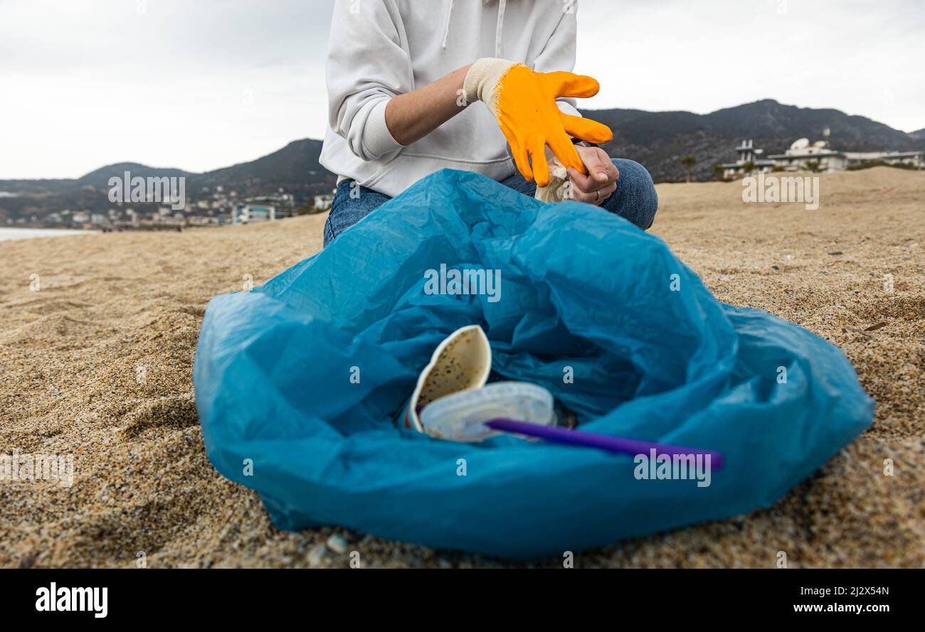 Eine Frau in Handschuhen mit einem speziellen blauen Beutel sammelt Müll im Sand entlang der Küste. Das Problem der Umweltverschmutzung. Müllabfuhr Stockfoto