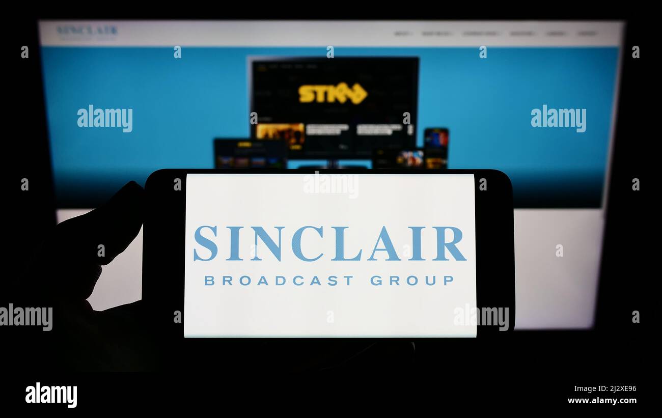 Person, die das Smartphone mit dem Logo der US-amerikanischen Firma Sinclair Broadcast Group Inc. (SBG) auf dem Bildschirm vor der Website hält. Konzentrieren Sie sich auf die Telefonanzeige. Stockfoto