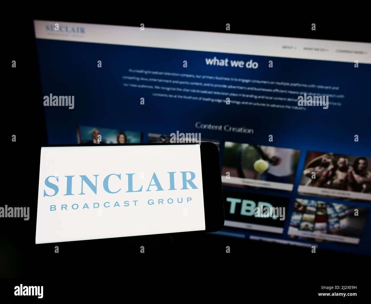 Person, die ein Mobiltelefon mit dem Logo des US-Unternehmens Sinclair Broadcast Group Inc. (SBG) auf dem Bildschirm vor der Webseite hält. Konzentrieren Sie sich auf die Telefonanzeige. Stockfoto
