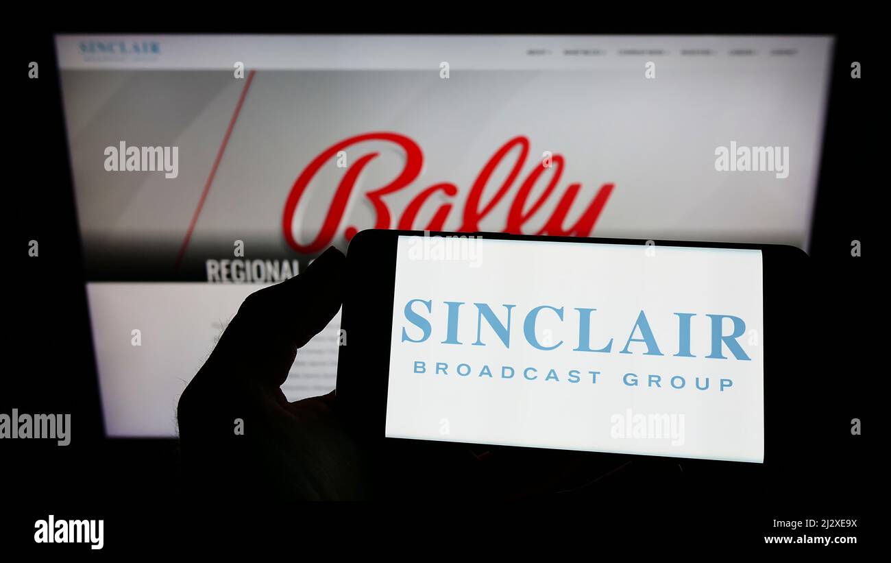 Person mit Mobiltelefon und Logo der amerikanischen Firma Sinclair Broadcast Group Inc. (SBG) auf dem Bildschirm vor der Webseite. Konzentrieren Sie sich auf die Telefonanzeige. Stockfoto