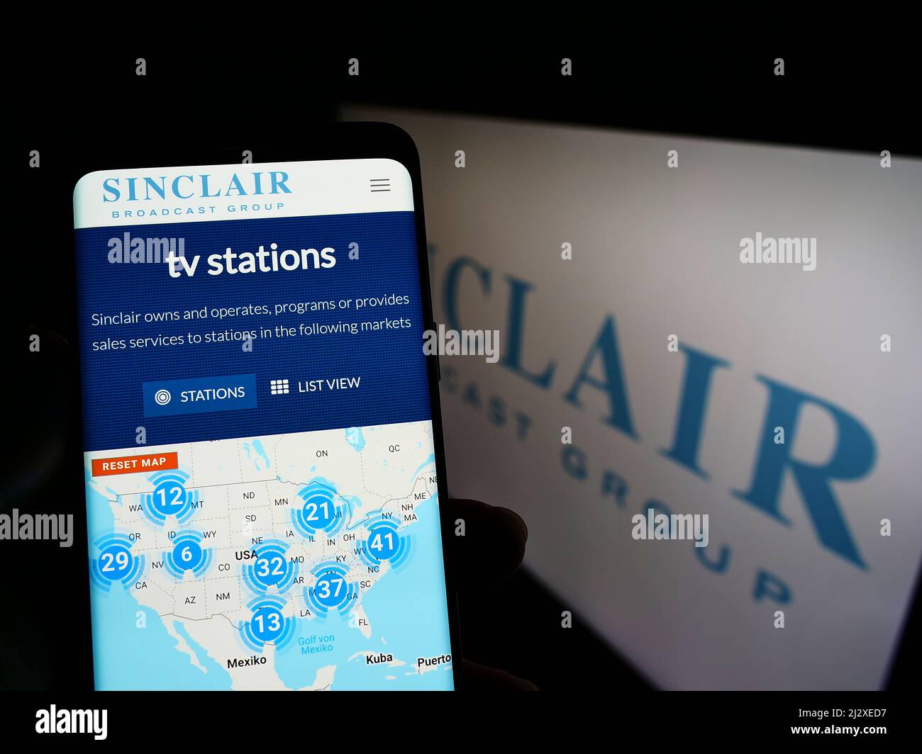 Person, die Mobiltelefon mit der Webseite des US-Unternehmens Sinclair Broadcast Group Inc. (SBG) auf dem Bildschirm mit Logo hält. Konzentrieren Sie sich auf die Mitte des Telefondisplays. Stockfoto