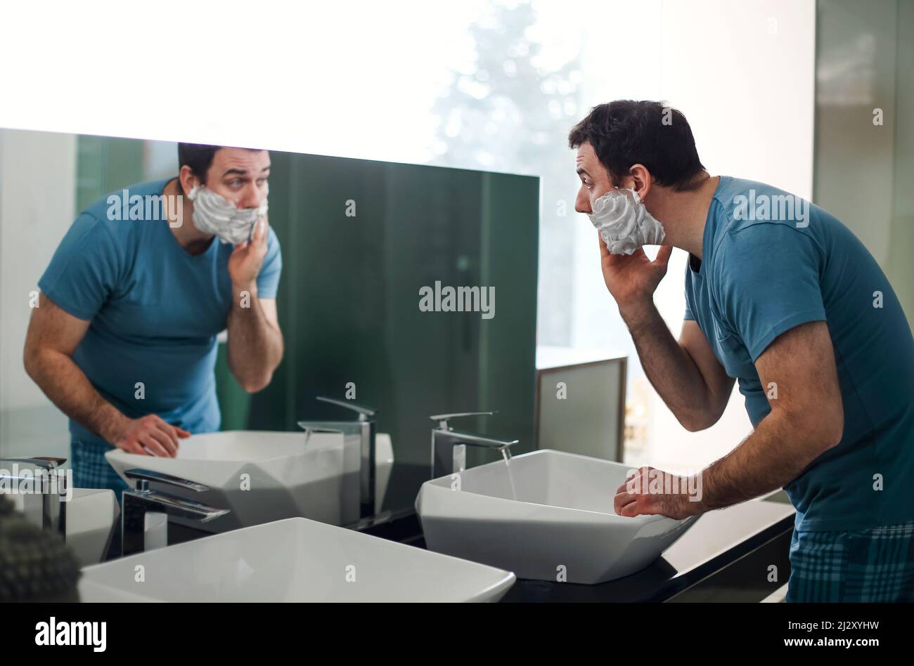 Hübscher Brünette Mann rasieren im Badezimmer. Er steht und schaut konzentriert auf den Spiegel. Mann, der Rasierschaum auf sein Gesicht aufbrachte Stockfoto