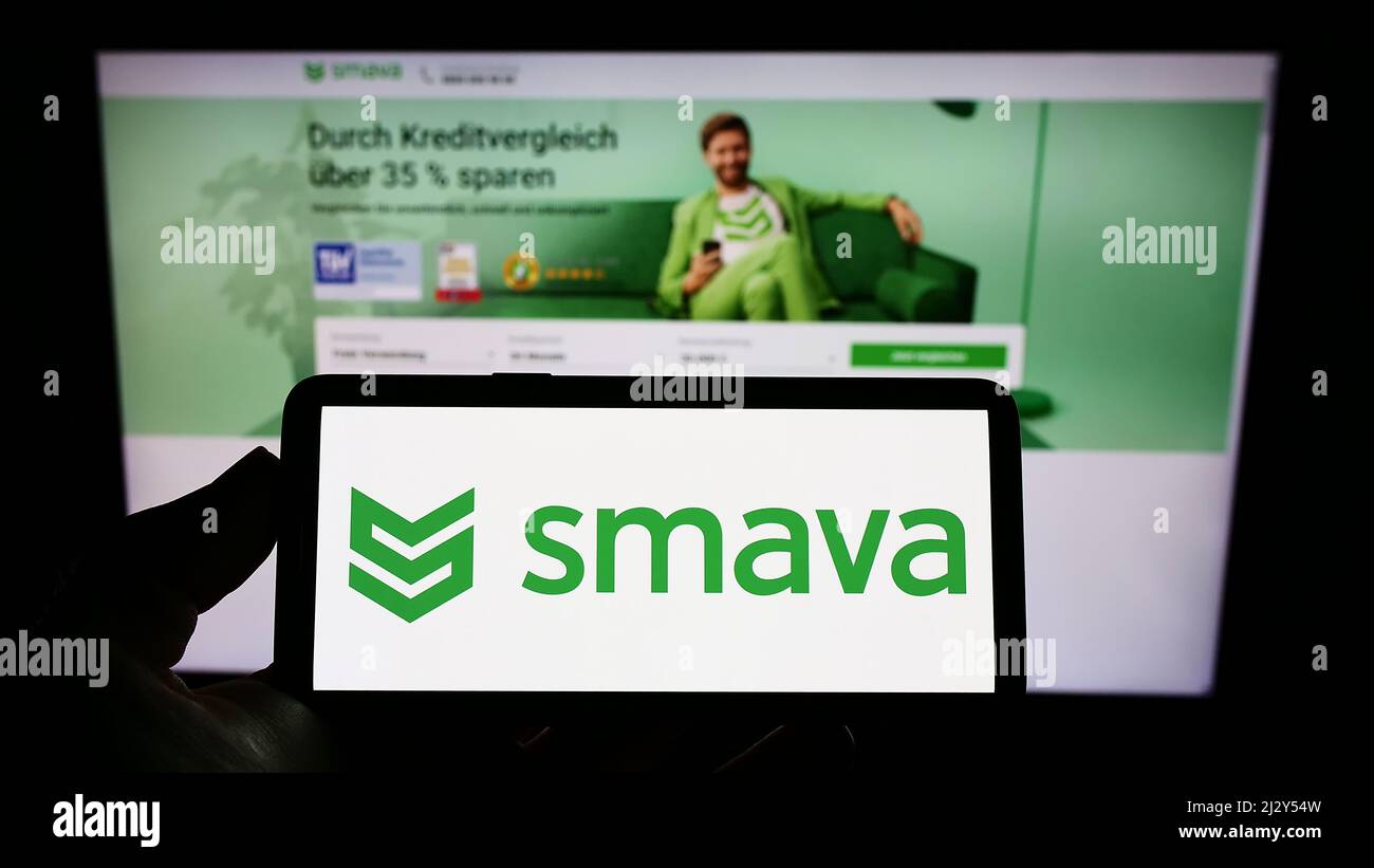 Person, die Mobiltelefon mit Logo der deutschen Kreditvergleichsgesellschaft smava GmbH auf dem Bildschirm vor der Business-Webseite hält. Konzentrieren Sie sich auf die Telefonanzeige. Stockfoto