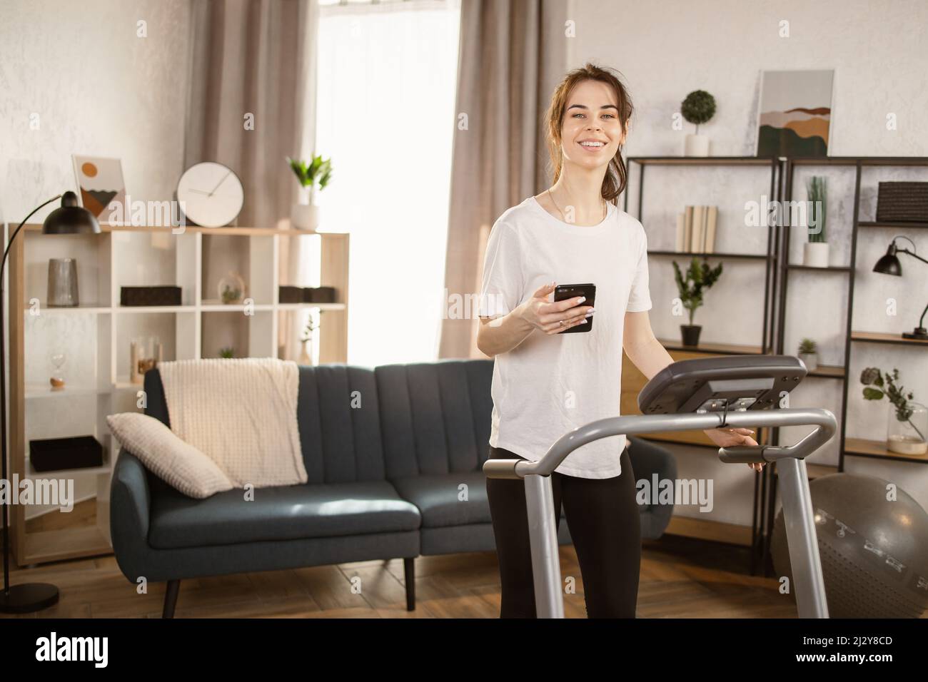 Vorderansicht einer sportlichen Frau, die ein Smartphone benutzt, während sie auf dem Laufband in schwarzen Sport-Leggings und einem weißen T-Shirt-Training zu Hause trainiert. Stockfoto