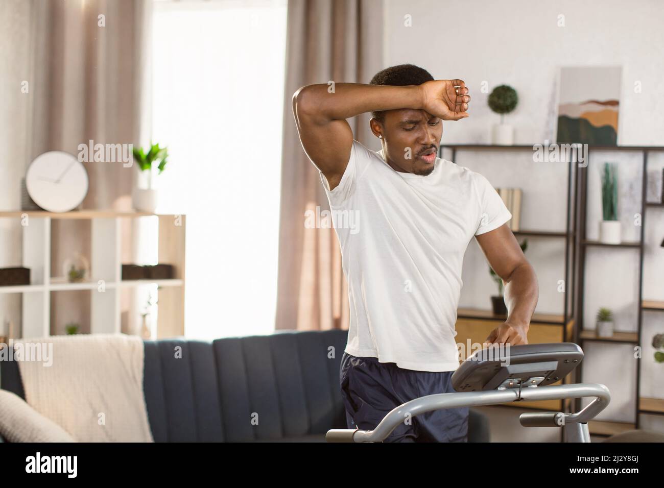 Junge sportliche afroamerikanische Männershorts und ein weißes T-Shirt trainieren zu Hause, machen Cardio-Übungen auf einem Laufband, während er Schweiß von der Stirn wischt. Konzept des Sports Gesundheit Aktion Ernährung Stockfoto