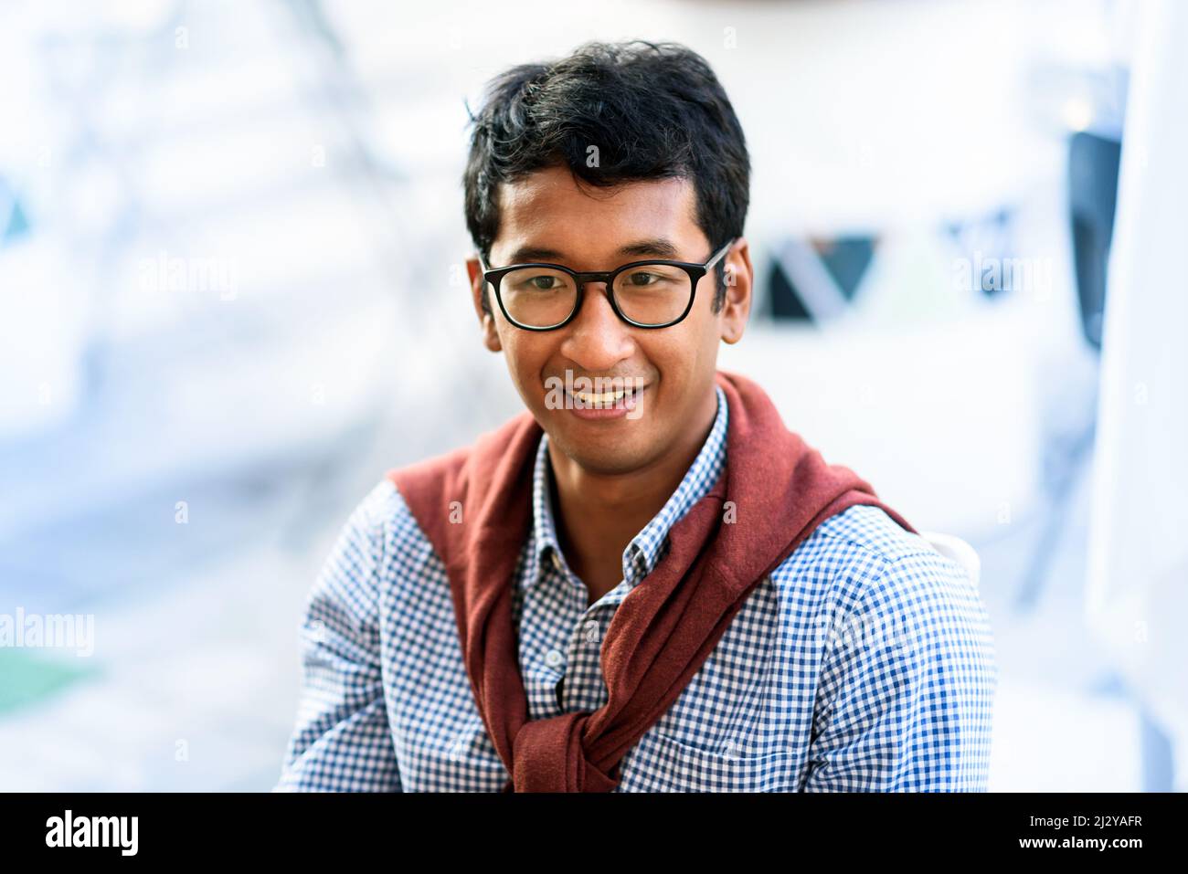 Porträt eines jungen asiatischen Mannes, der eine Brille im Freien trägt, mit einem Pullover, der über seine Schultern vor einem Hintergrund mit hohem Schlüsselüberstand drapiert ist Stockfoto