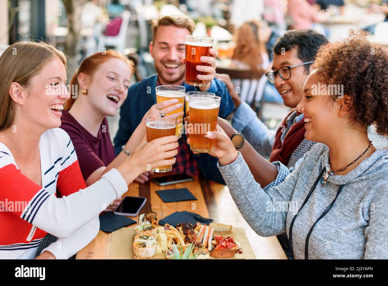 Eine Gruppe von positiven multirassischen Freunden klirren Biergläser an, während sie mit Snacks am Tisch sitzen, während sie ihren Urlaub auf der Terrasse des Pubs im Freien feiern Stockfoto