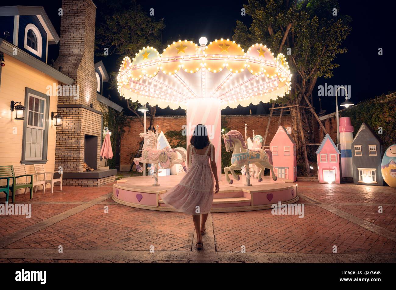 Rückansicht einer asiatischen Frau in Kleid, die in der Nacht zum beleuchteten Karussell im Vergnügungspark läuft Stockfoto