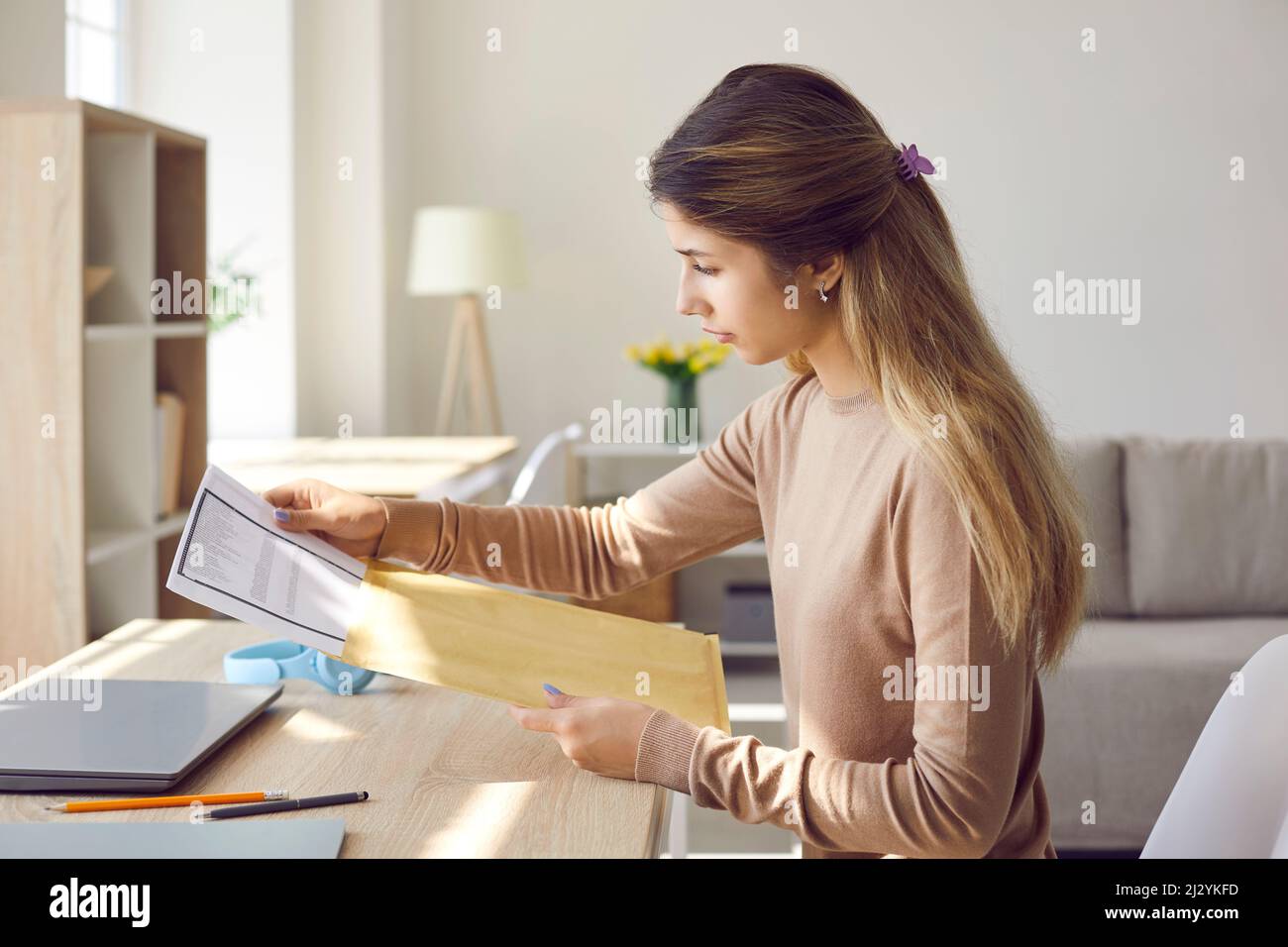 Studentin nimmt Brief aus dem Umschlag, während sie zu Hause am Schreibtisch mit Laptop sitzt Stockfoto