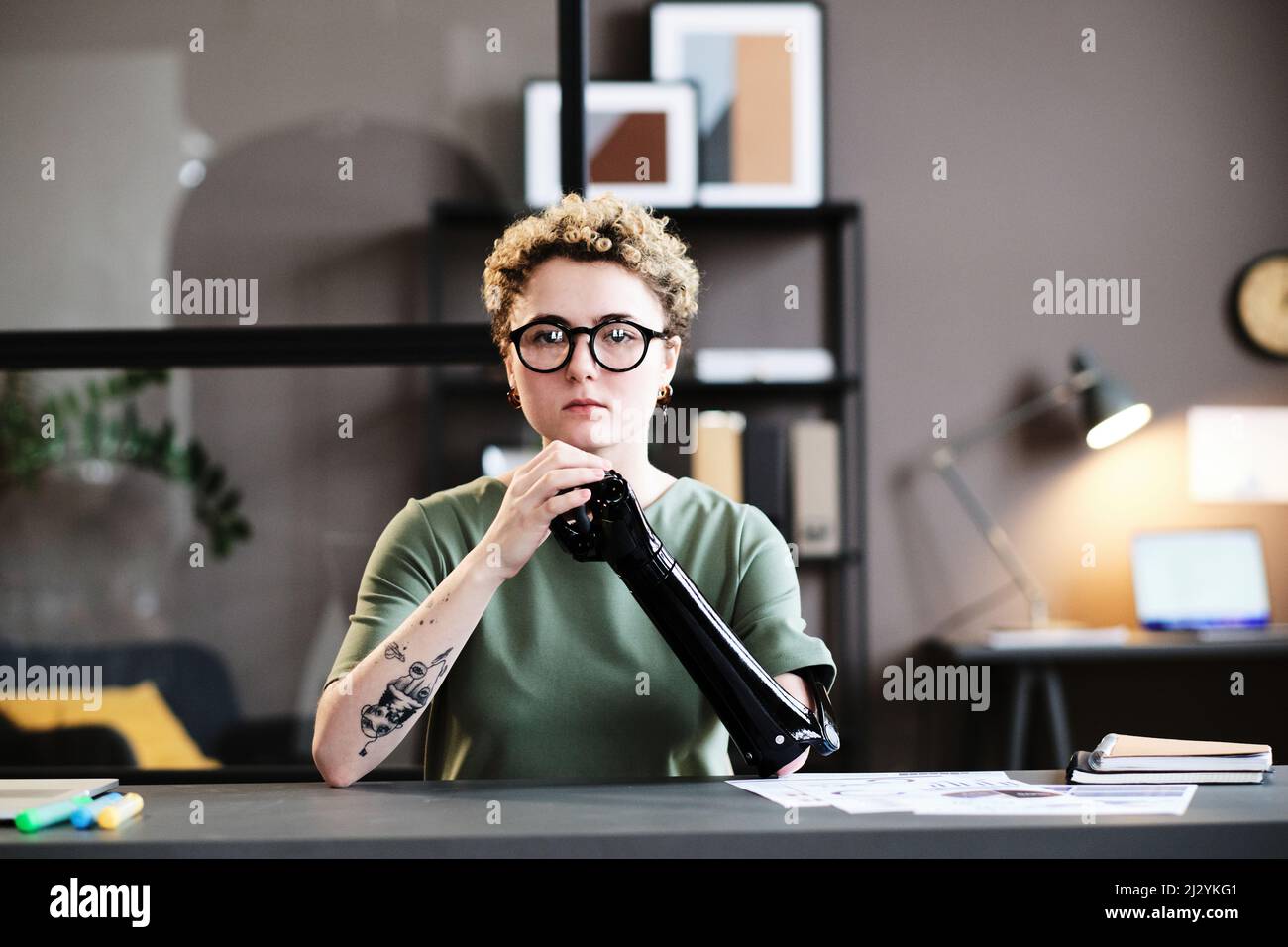 Porträt einer jungen Frau in einer Brille mit Prothesenarm, die auf die Kamera schaut, während sie am Schreibtisch sitzt Stockfoto