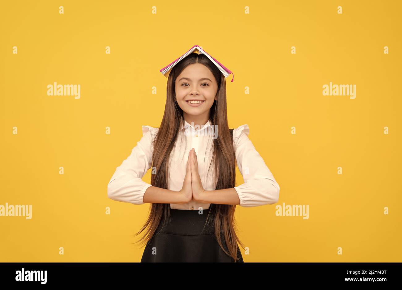 Glückliches Teenager-Mädchen in Schuluniform mit Buch auf Kopf meditieren mit Handbewegung, Ruhe bewahren Stockfoto
