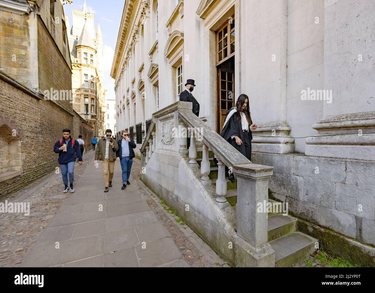 Town and Gown, im Gegensatz zu Großbritannien; eine Studentin der Cambridge University steigt an ihrem Abschlusstag aus dem Senatshaus auf, während einheimische Männer an Cambridge, Großbritannien, vorbeigehen Stockfoto