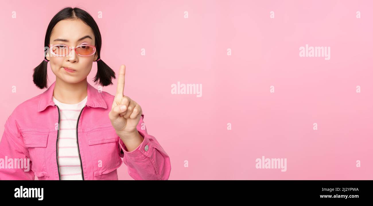 Bild von schweren, stilvollen asiatischen Mädchen in Sonnenbrille, zeigt Stopp, Geste verbieten, Tabuschild, nein sagen, Stehen auf rosa Hintergrund Stockfoto