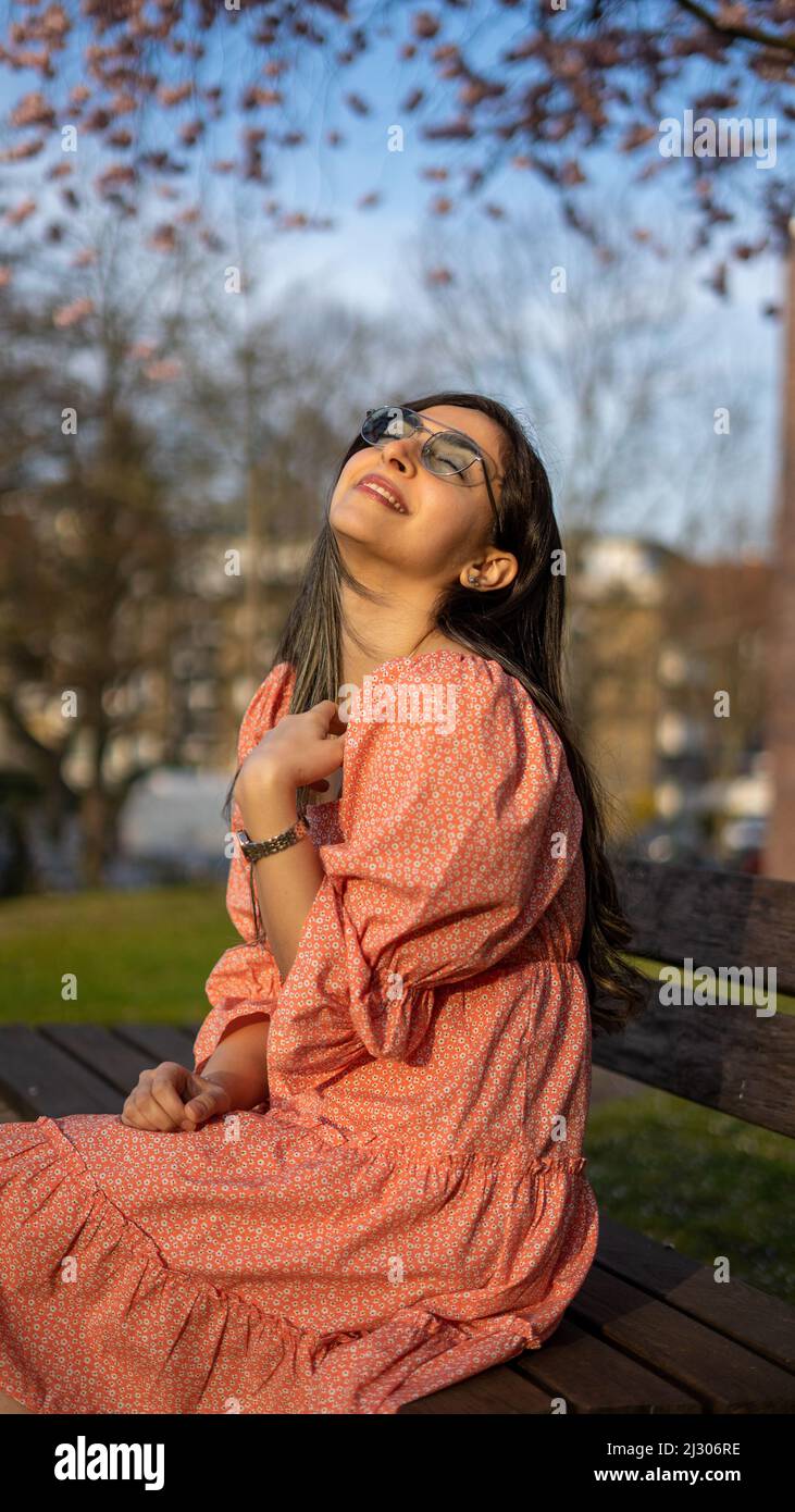Schöne iranische junge Frau mit Sonnenbrille und buntem Kleid im Frühjahr mit einem Kirschblütenbaum auf dem Hintergrund Stockfoto