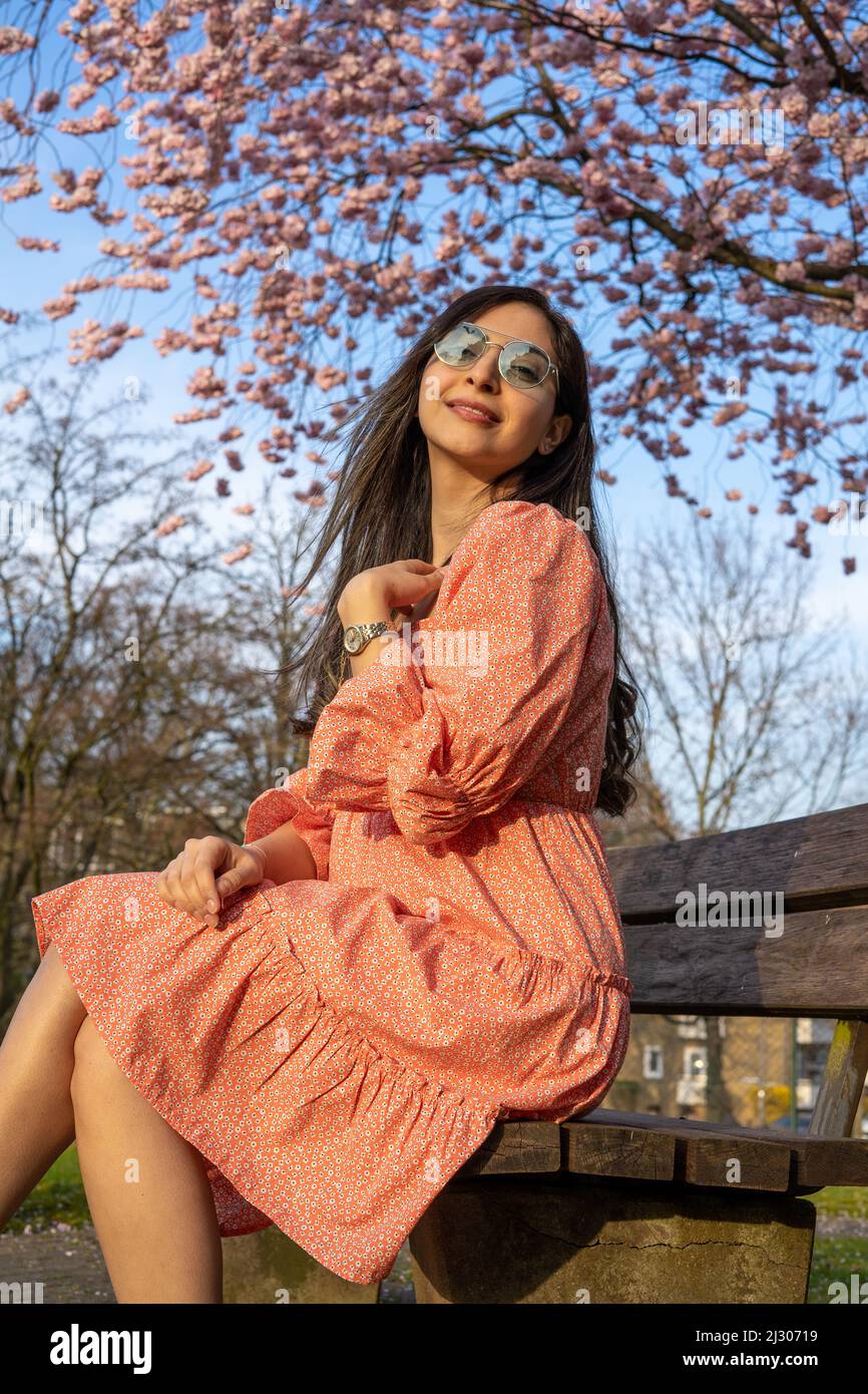 Schöne iranische junge Frau mit Sonnenbrille und buntem Kleid im Frühjahr mit einem Kirschblütenbaum auf dem Hintergrund Stockfoto
