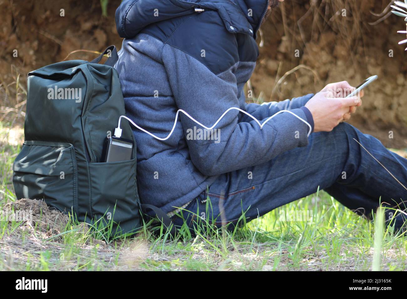 Mann sitzt auf dem Boden und hält ein Smartphone in den Händen und lädt es vor dem Hintergrund eines Naturschauers mit einer schwarzen Powerbank auf. Tragbares Reiseladegerät. Stockfoto