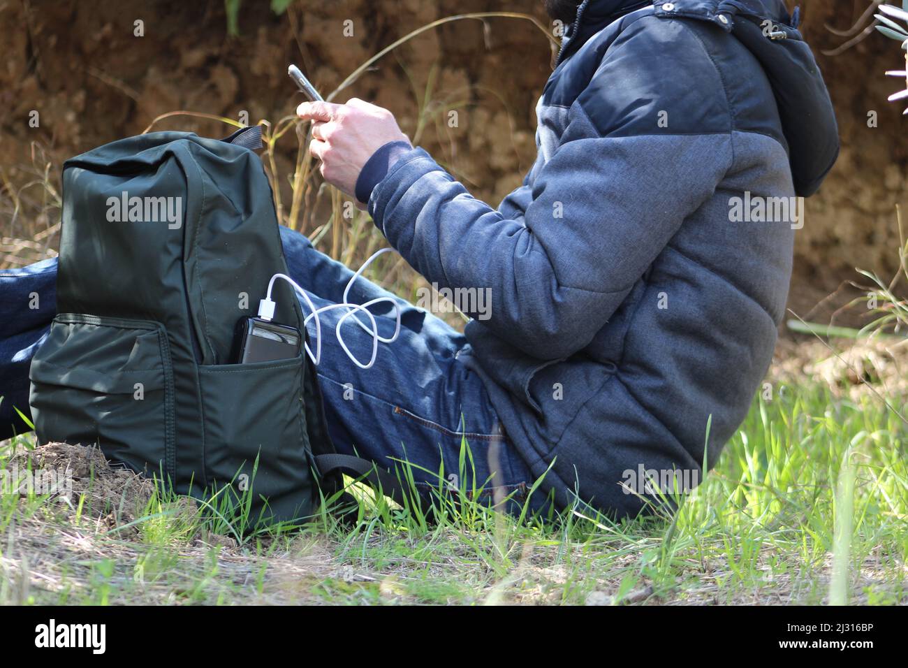 Mann sitzt auf dem Boden und hält ein Smartphone in den Händen und lädt es vor dem Hintergrund eines Naturschauers mit einer schwarzen Powerbank auf. Tragbares Reiseladegerät. Stockfoto
