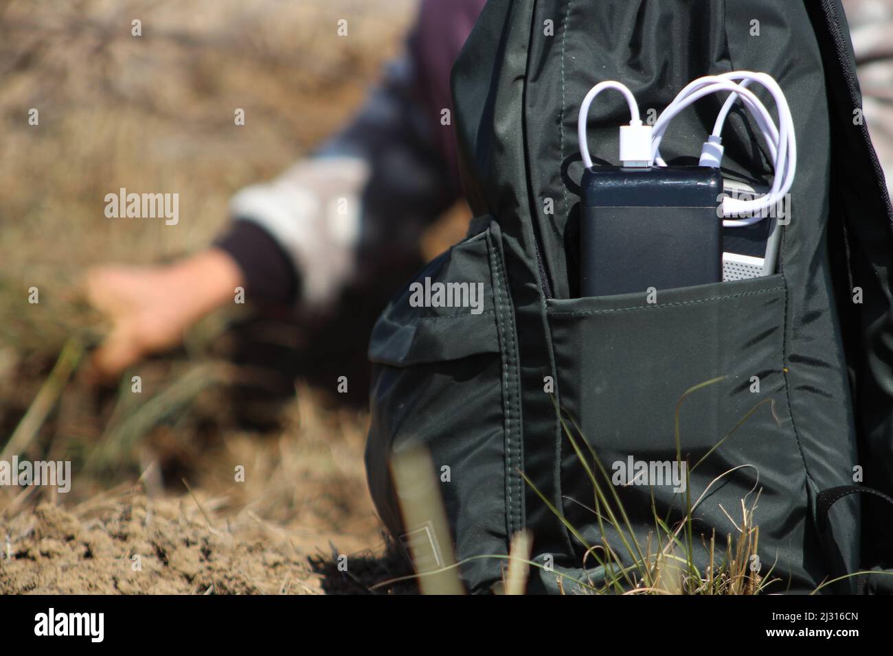 Smartphone mit Powerbank im Rucksack aufgeladen. Eine Frau im Hintergrund zündet in der Natur ein Lagerfeuer an. Stockfoto