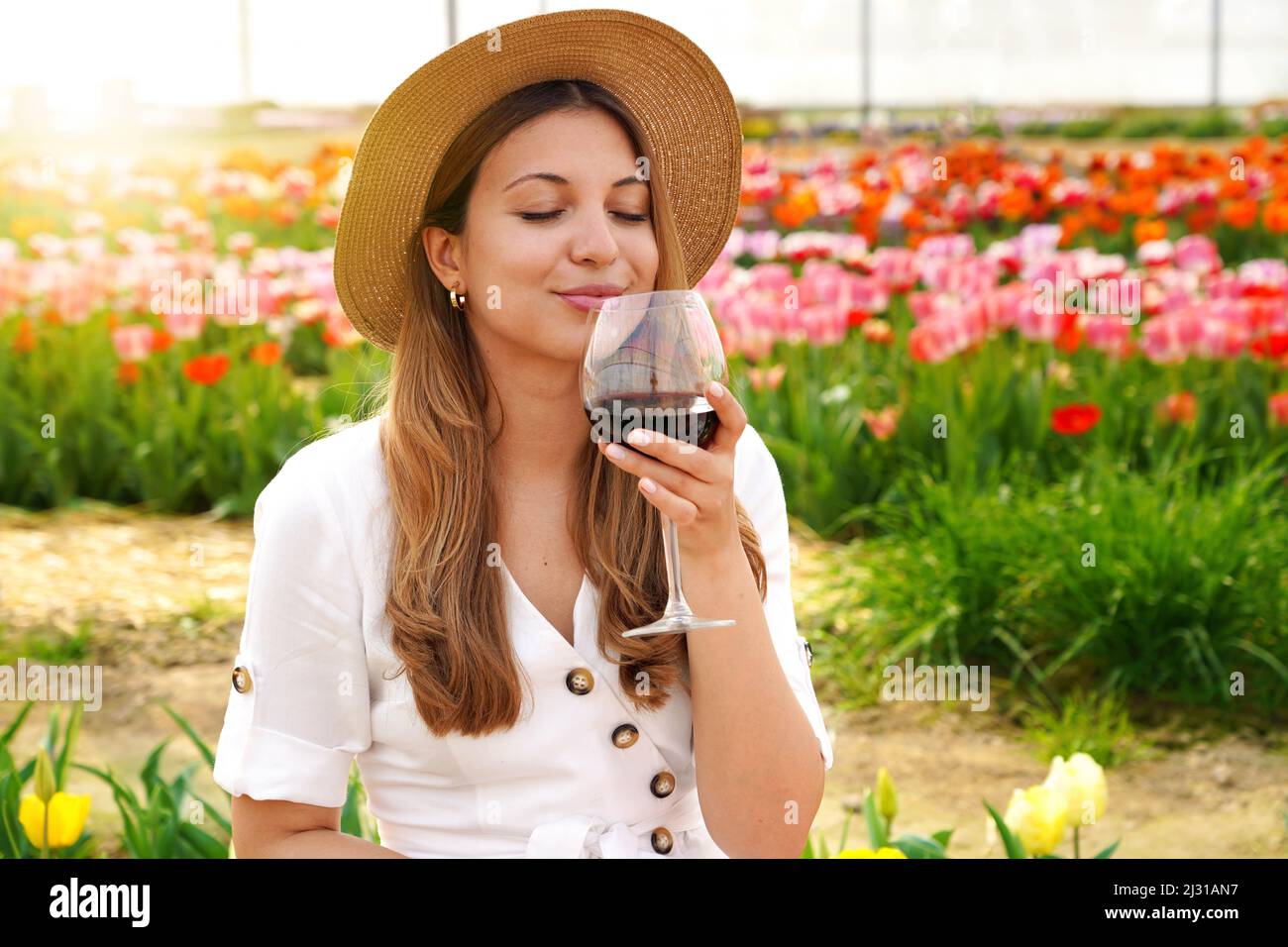 Schöne Frau riecht kostbaren Rotwein mit geschlossenen Augen im Garten mit blühenden Blumen auf dem Hintergrund Stockfoto