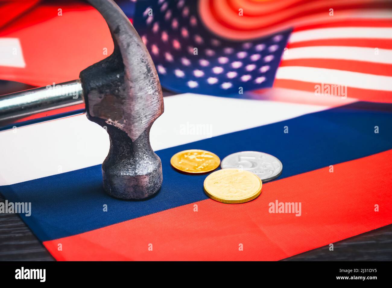 Russische Rubel-Münzen werden mit einem Metallhammer vor dem Hintergrund der russischen und der US-amerikanischen Flagge zerschlagen Stockfoto