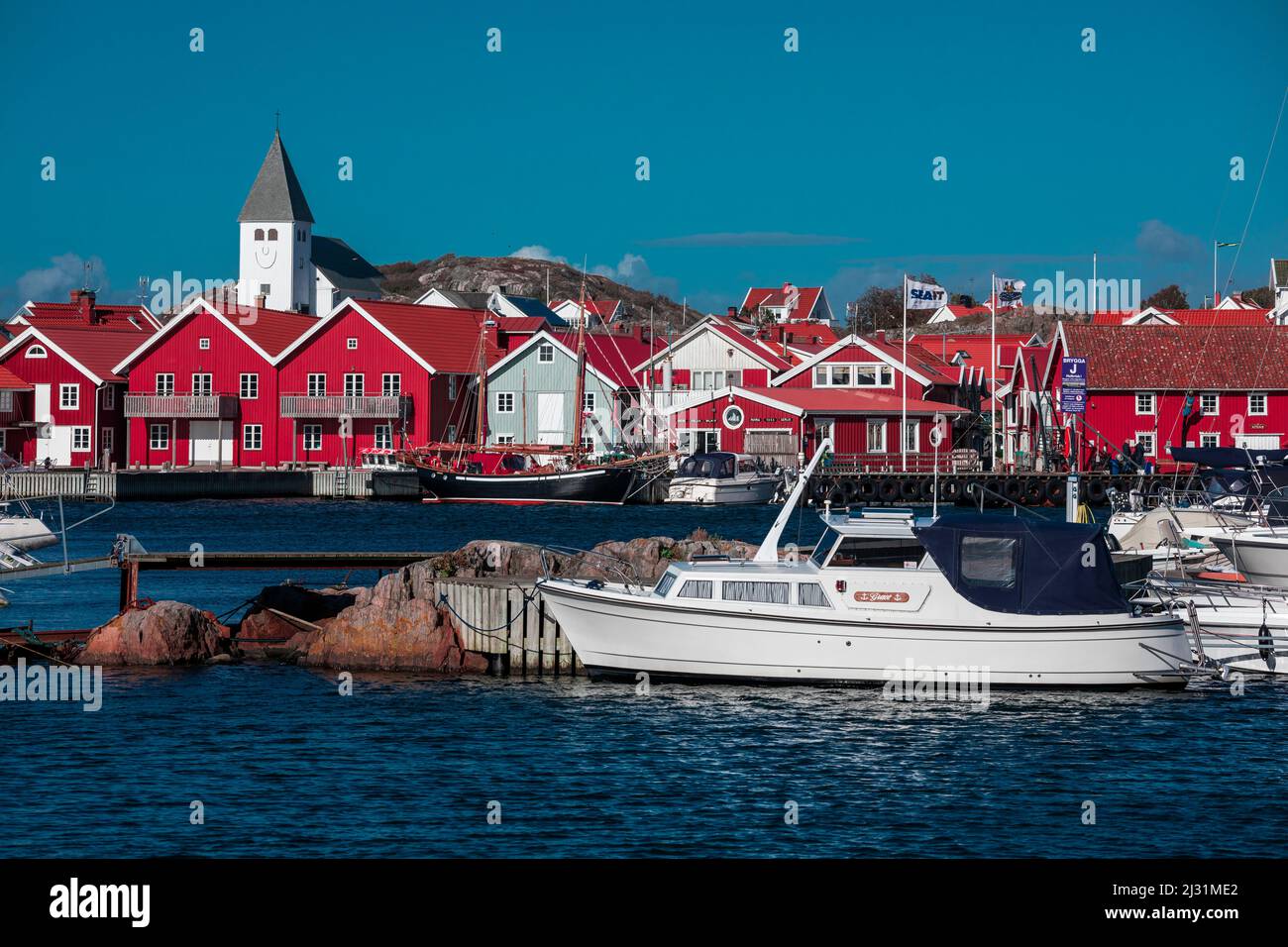 Rote Häuser mit Kirche und Booten im Dorf Skärhamn auf der Schäreninsel Tjörn an der Westküste Schwedens, blauer Himmel mit Sonne Stockfoto