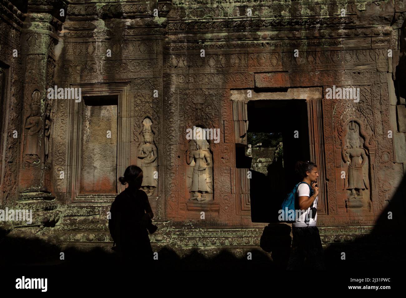 Weibliche Besucher auf einem Fußweg vor einer mit apsara-Schnitzereien geschmückten Mauer im Ta Prohm, einem alten Kloster und Tempel in Siem Reap, Kambodscha. Stockfoto