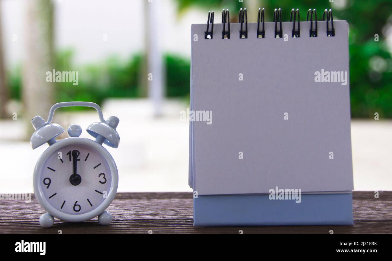 Wecker, der auf 12 Uhr zeigt, mit leerem Notizblock. Verschwommener Parkhintergrund. Konzept für den Kopierraum Stockfoto