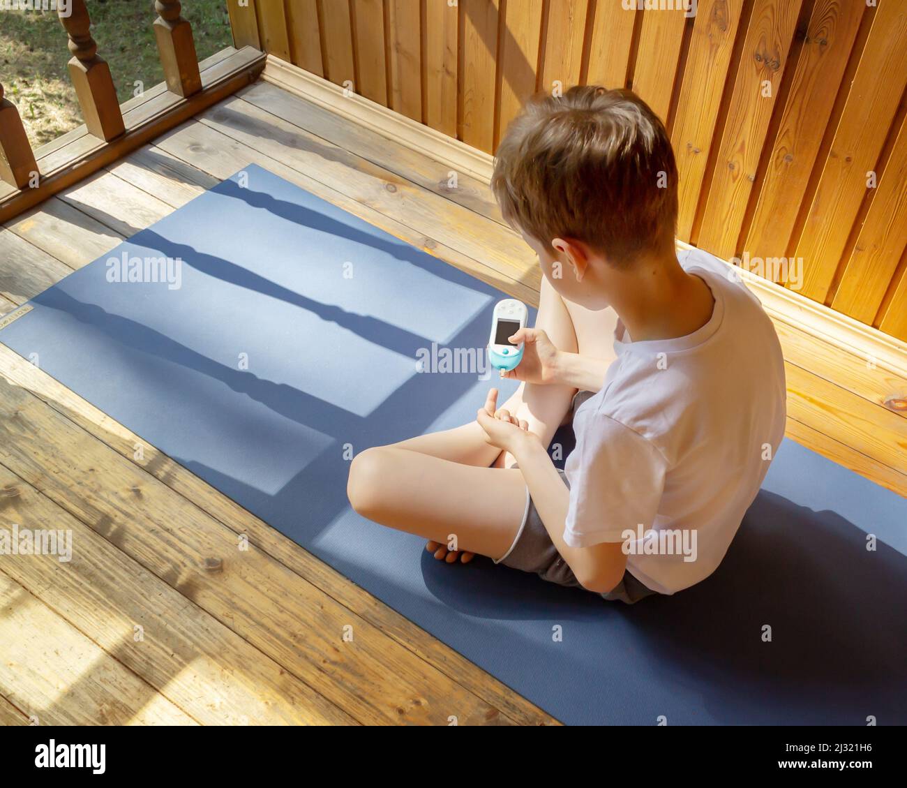Konzept des Lebens eines Kindes mit Diabetes und glykämischer Kontrolle. Sommerurlaub: Ein Junge sitzt auf der Sportmatte im Freien und misst den Zuckerspiegel Stockfoto