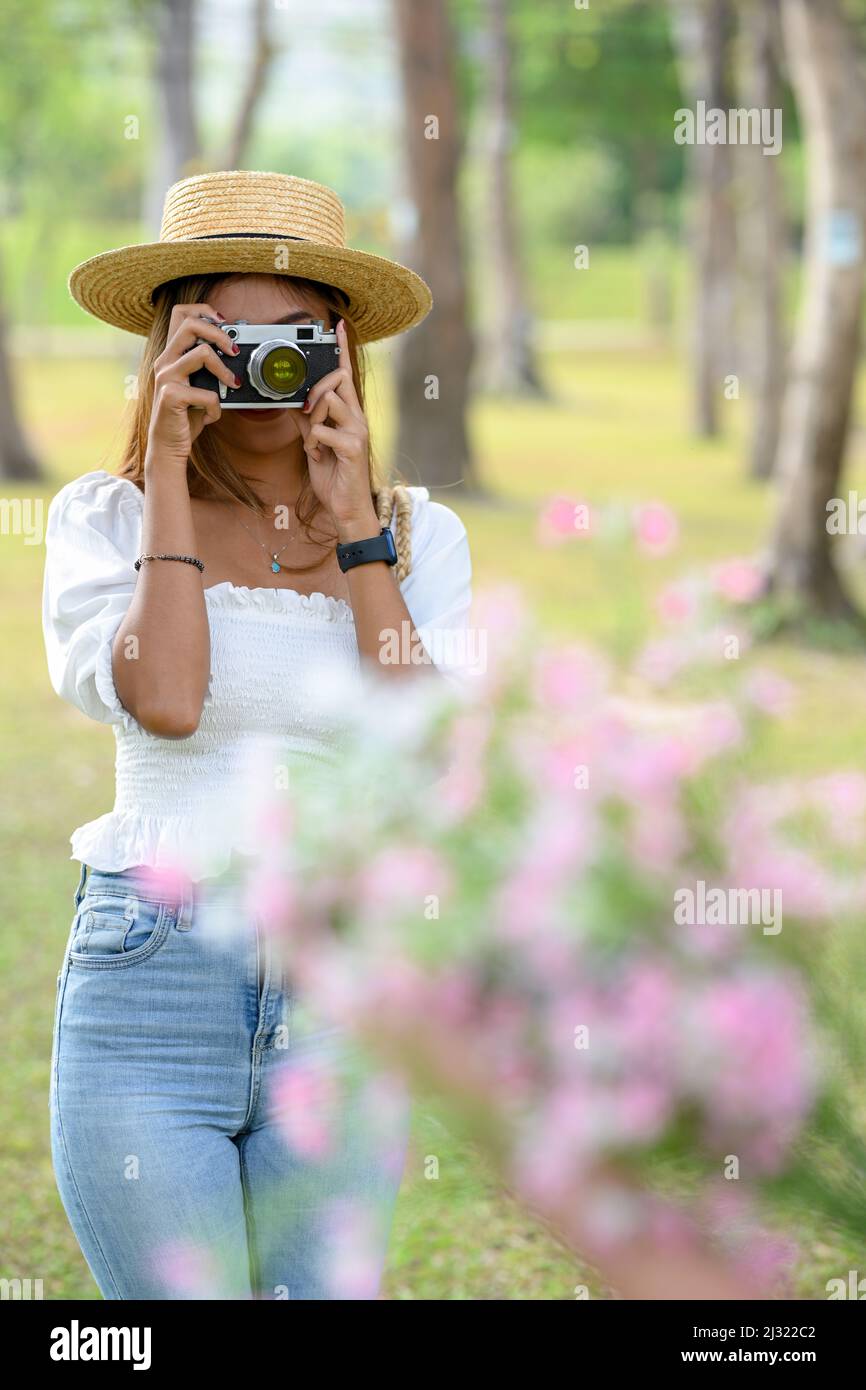 Porträt einer attraktiven asiatischen Frau in kausalem Outfit und Strohhut, die mit einer Retro-Kamera im Garten ein Blumenbild macht. Stockfoto