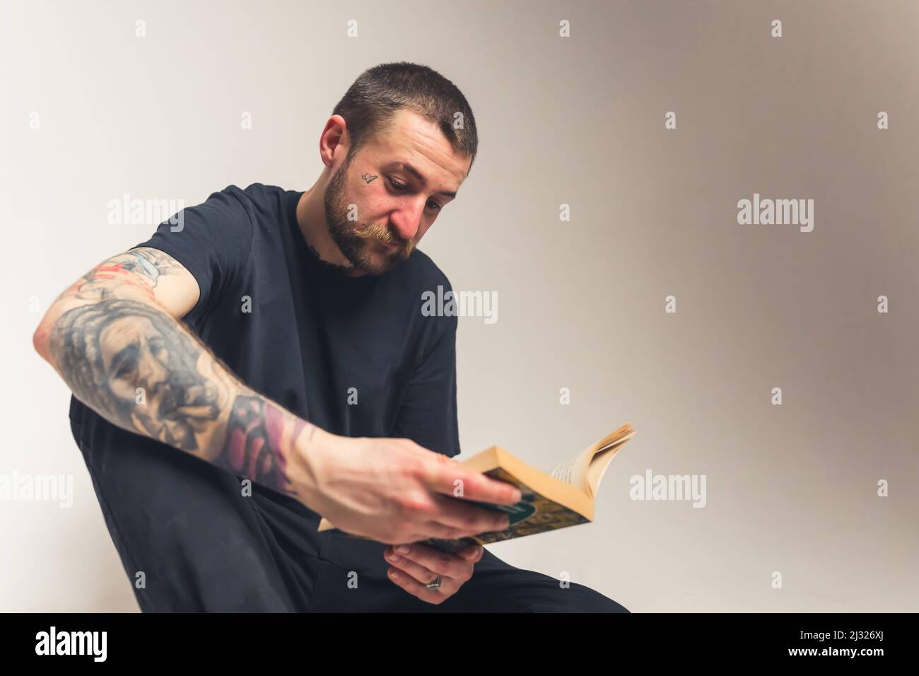 Hübscher bärtiger Kaukasischer tätowierte Mann, der auf dem Boden sitzt und die Heilige Bibel liest - Faith Konzept isoliert auf hellem Hintergrund mittlere Ganzaufnahme Studioaufnahme. Hochwertige Fotos Stockfoto