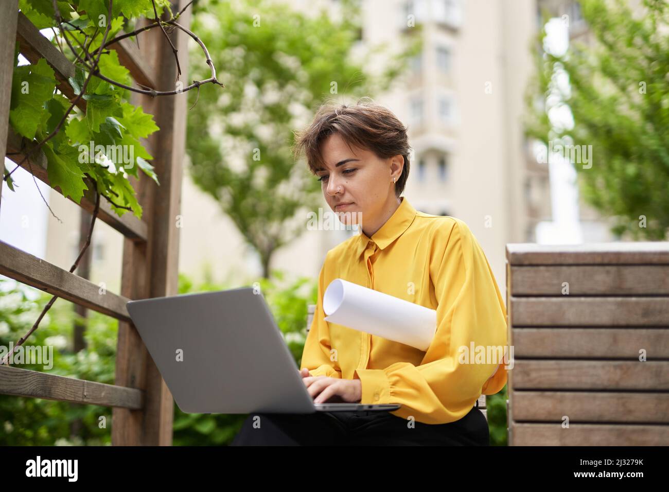 Erfolgreiche lächelnde Geschäftsfrau in gelber Bluse sitzt auf der Bank im Freien im Park mit Plänen mit Laptop arbeiten oder Online-Meeting. Business, Finanzindustrie oder Immobilienkonzept Stockfoto