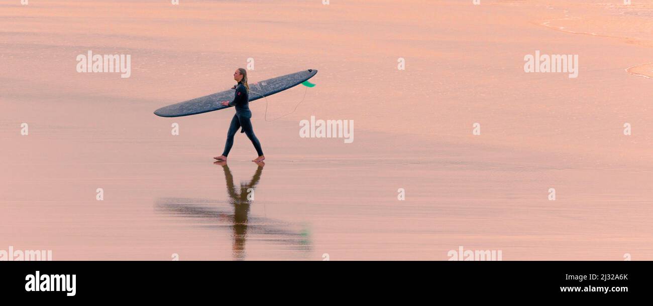 Ein Panoramabild des warmen Abendlichts über einer Surferin, die sein Surfbrett trägt und nach einer Surfstunde in Newquay in Fistral Beach spazierengeht Stockfoto