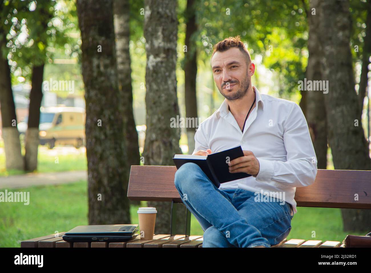 Ein lächelnder Mann sitzt auf einer Bank, macht Pläne, schreibt in ein Notizbuch. Ein junger Mann auf einem Hintergrund von grünen Bäumen, ein heißer sonniger Sommertag. Warmes, weiches Licht, Nahaufnahme. Stockfoto
