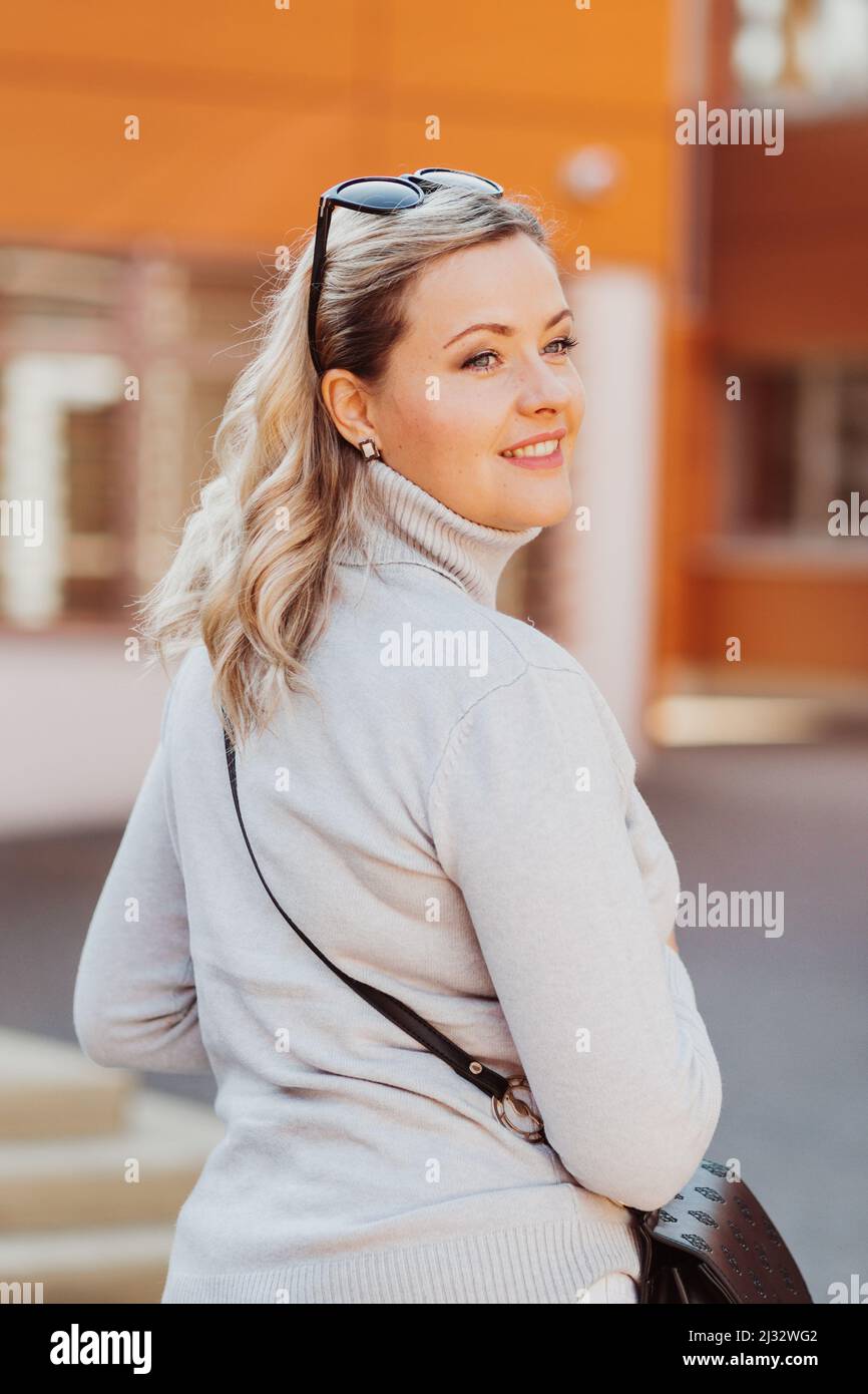 Vertikale lächelnde Frau mit Sonnenbrille, schwarzer Umhängetasche, lässiger weißer Pullover, Umdrehen. Sonnenorange. Nahaufnahme Stockfoto