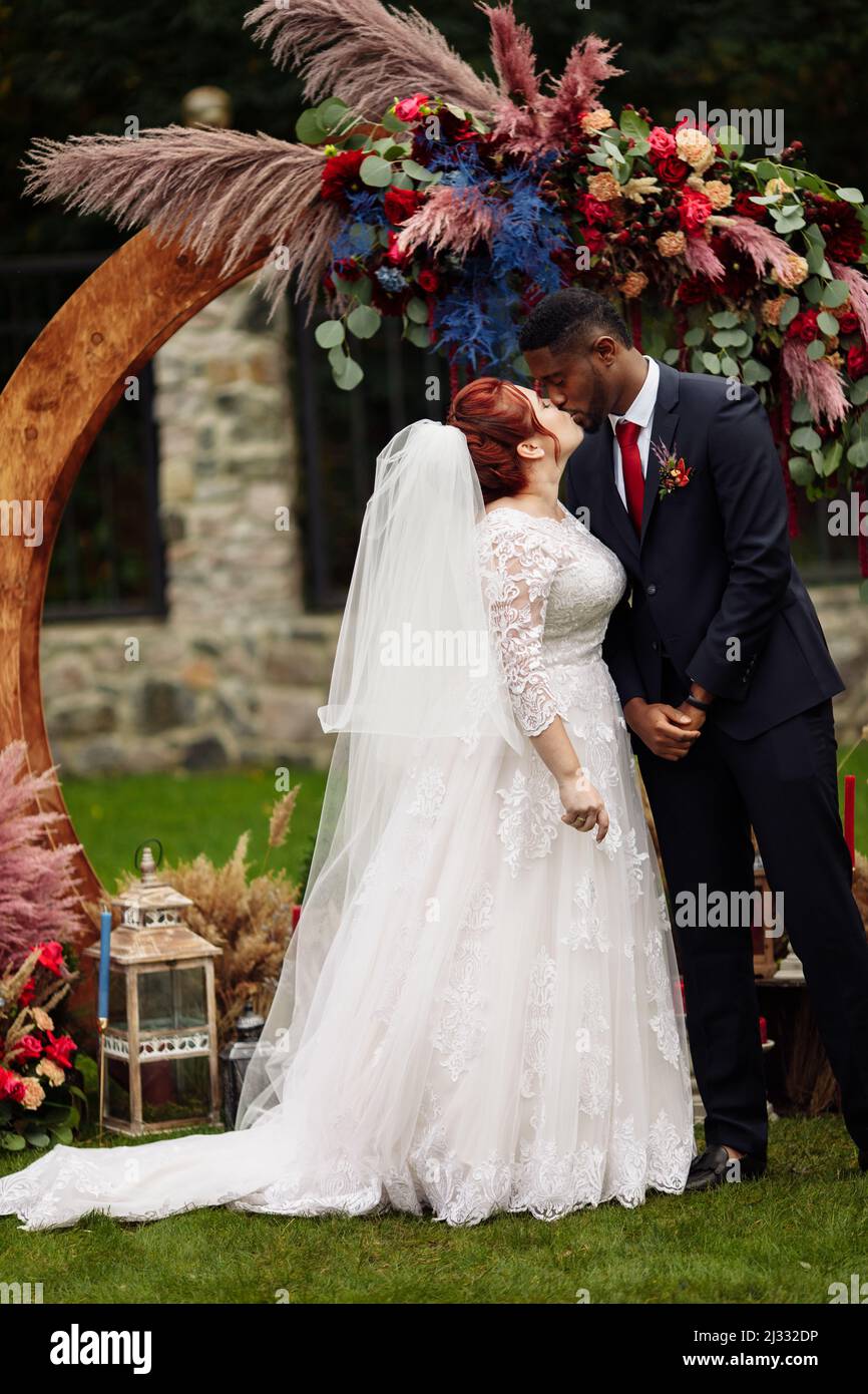 Charmanter afroamerikanischer Bräutigam mit wunderschönen weißen Braut stehen in der Nähe dekorierten Bogen, schöne Frau küssen schönen Mann auf Hochzeitszeremonie, glücklich gemischt r Stockfoto